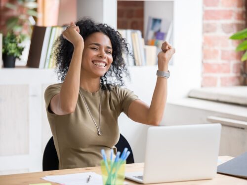 A young woman seated at a computer in a modern office setting raises her hands in celebration