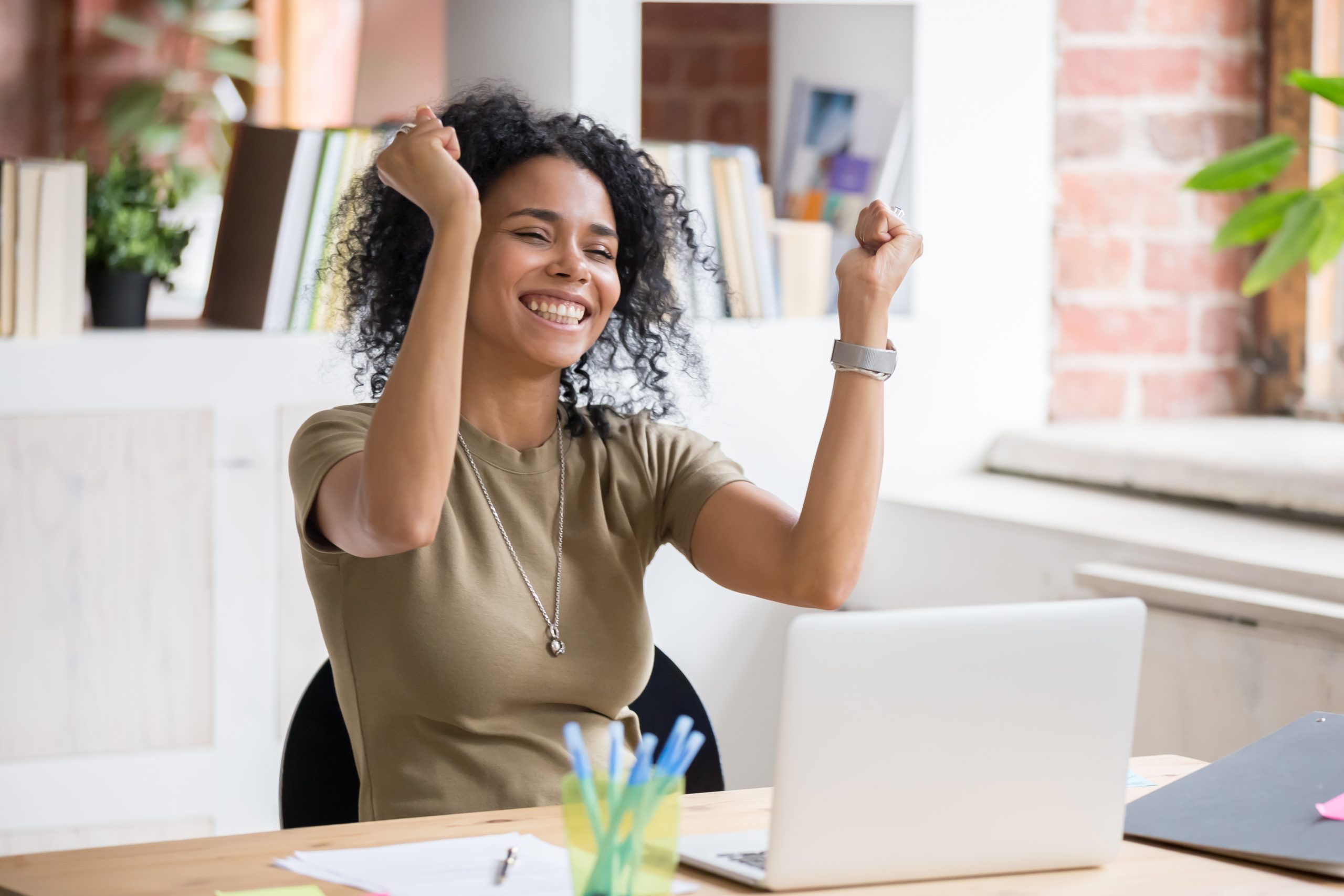 A young woman seated at a computer in a modern office setting raises her hands in celebration